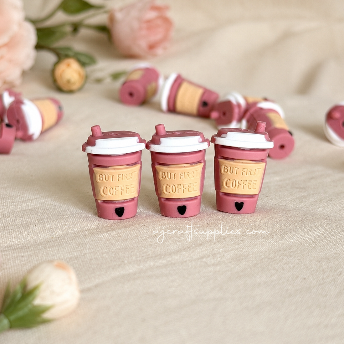 Three small pink and beige coffee cup charms with the words 'BUT FIRST COFFEE' embossed on the cups, arranged on a beige fabric surface with soft pink flowers in the background.